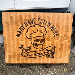 Wooden cutting board with skull and chef hat design and text 'Many have eaten here, few have died' on a kitchen counter.